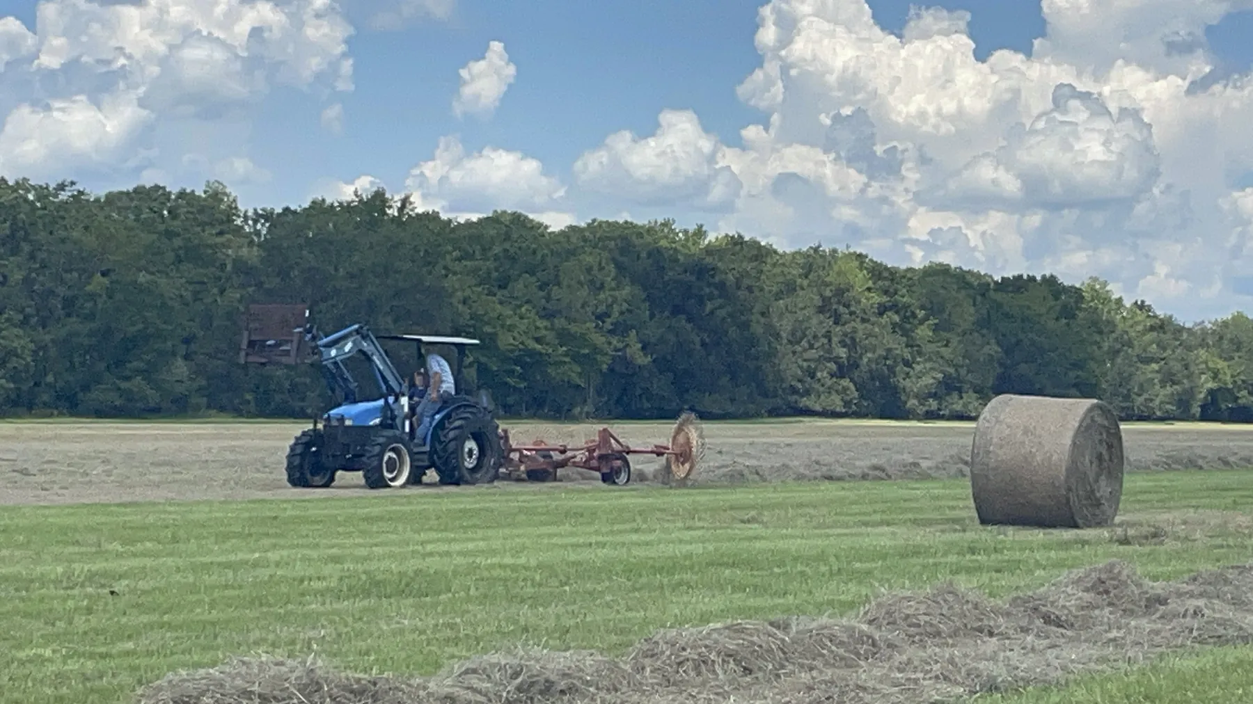A blue tractor raking hay into rows next to a round bale